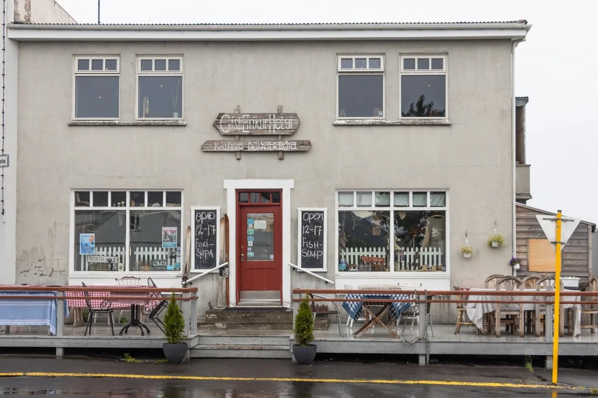 Façade d'un café-hôtel gris avec porte rouge et terrasse en bois