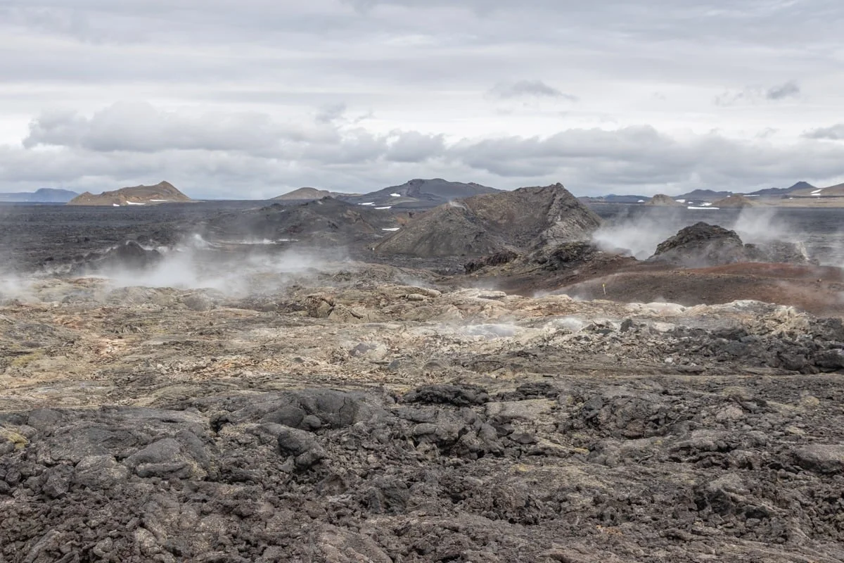 Champ de lave noire avec fumerolles et cratères volcaniques sous ciel nuageux