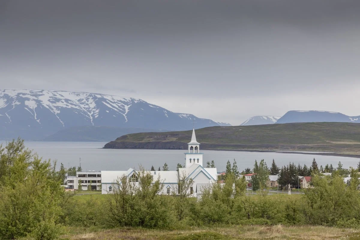 Église blanche avec clocher entourée d'arbres face à un fjord et des montagnes enneigées