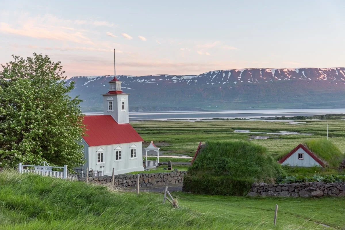 Petite église blanche à toit rouge entourée de prairies verdoyantes et montagnes enneigées
