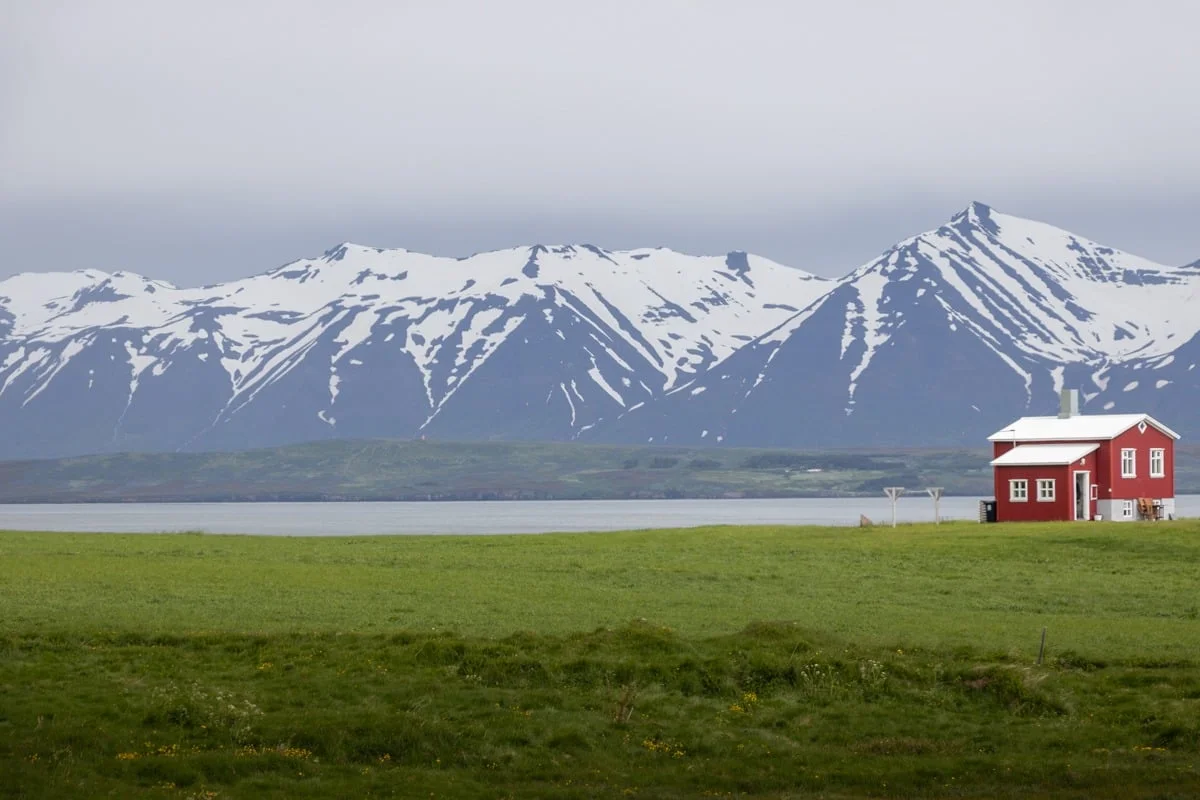 Petite maison rouge dans une prairie verte face à des montagnes enneigées
