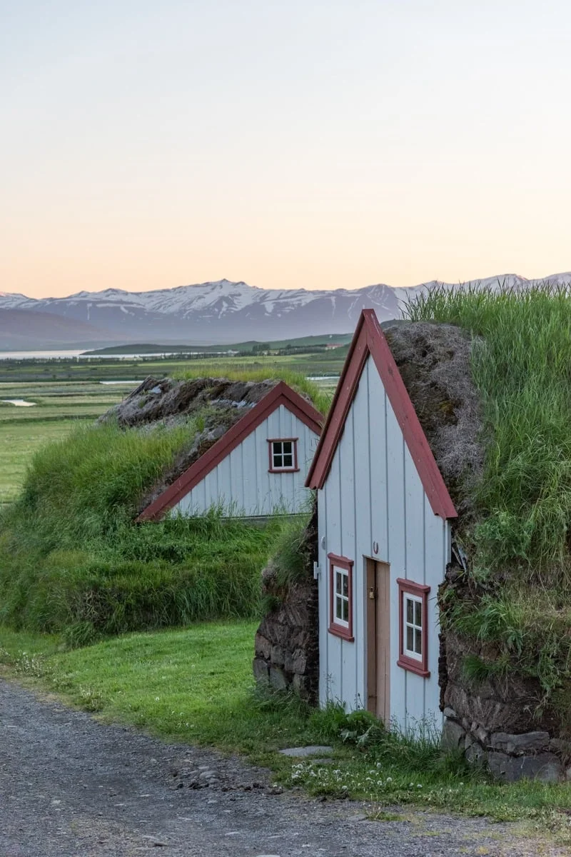 Deux petites maisons en tourbe aux façades blanches et encadrements bordeaux, prairies et montagnes enneigées en arrière-plan