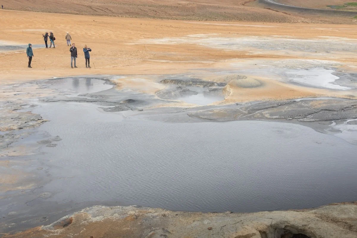 Mare de boue grise fumante entourée de sol ocre avec des visiteurs au fond