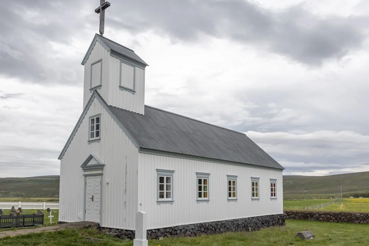 Petite église en bois blanc avec clocher et croix, entourée de verdure