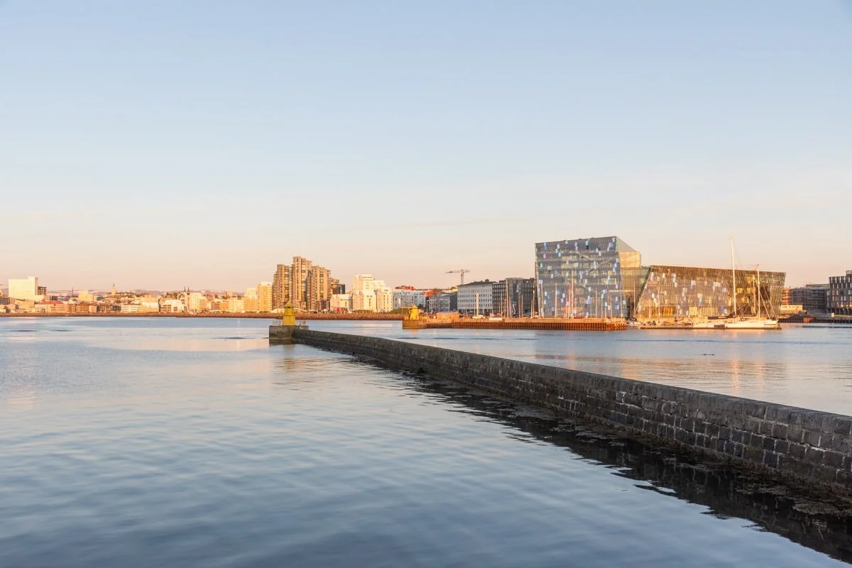 Jetée en pierre longeant le port de Reykjavik avec bâtiment en verre du Harpa au coucher du soleil