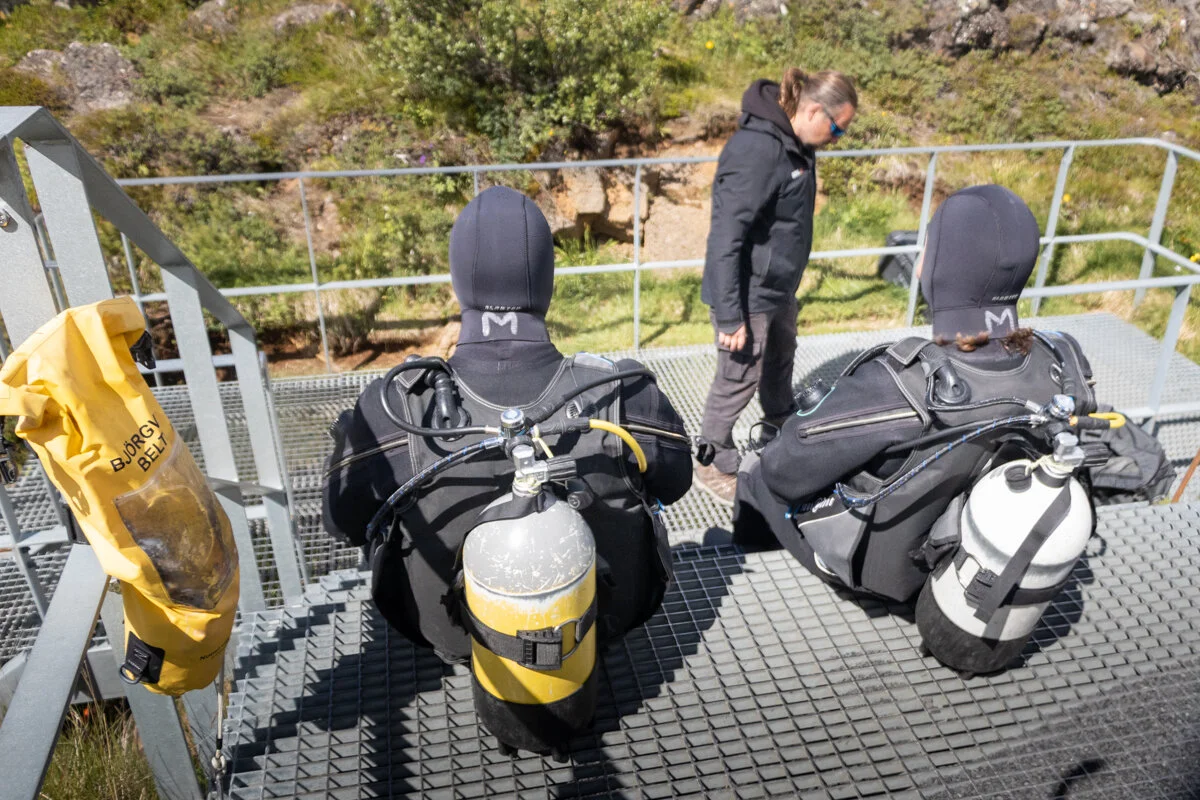 deux personnes assises, de dos, en combinaison et avec des bouteilles de plongée sur le dos